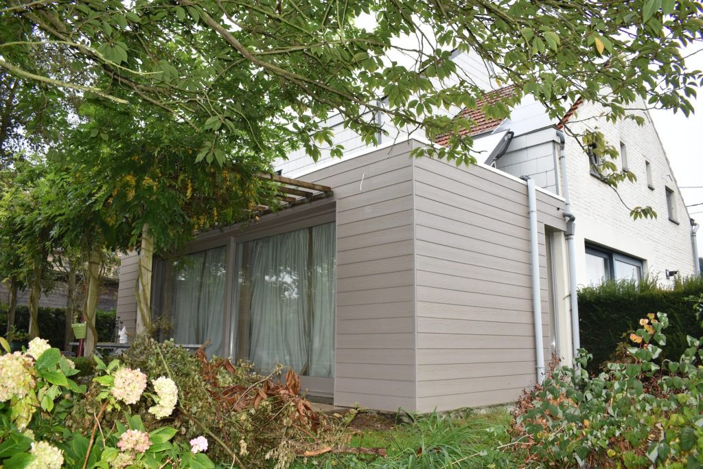 Moderne aanbouw met horizontale houtlook sidings in een warme taupe tint, naast een witte woning in een tuin.